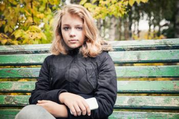 Beautiful Caucasian blond teenage girl in black jacket sitting on green park bench with smartphone in hands