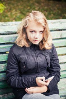 Beautiful Caucasian blond teenage girl in black jacket sitting on old green park bench with smartphone in hands