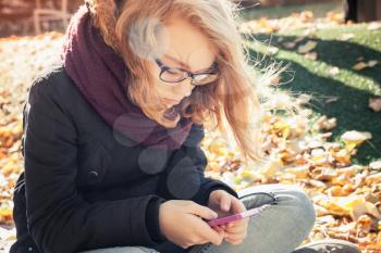 Cute Caucasian blond teenage girl in jeans and black jacket sitting in park and using smartphone, outdoor autumn portrait