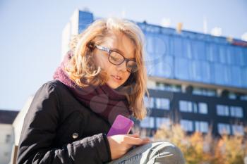 Beautiful Caucasian blond teenage girl in glasses sitting in city park and holds smartphone in hands