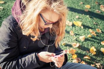 Caucasian blond teenage girl in glasses sitting in autumnal park and using smartphone