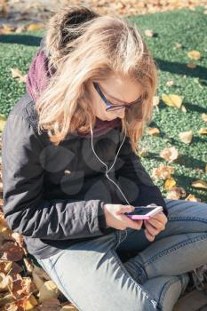 Caucasian blond teenage girl in jeans and black jacket sitting in park and using smartphone, outdoor autumn portrait, vintage style tonal correction photo filter