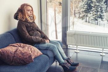 Beautiful blond Caucasian teenage girl in warm clothes sitting on blue sofa near the window in hallway