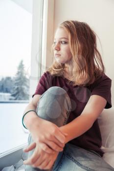 Blond teenage girl sits near winter window on a windowsill and looks outside