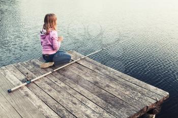 Little blond Caucasian girl sitting on a wooden pier with fishing rod, vintage stylized photo with warm tonal correction filter effect