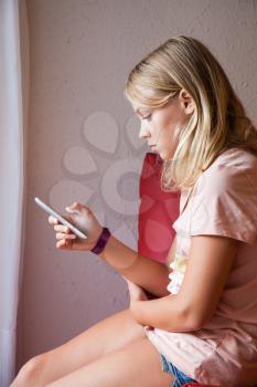 Cute Caucasian blond teenage girl in pink t-shirt using smartphone for messaging, indoor profile portrait