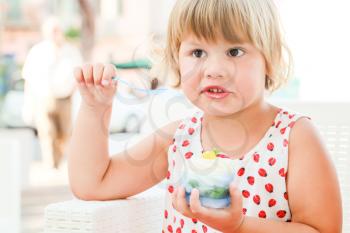 Cute blond Caucasian baby girl eats frozen yogurt with ice cream and fruits, closeup outdoor portrait with natural high key light