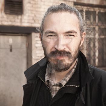 Outdoor closeup square portrait of young bearded Asian man in black on grungy building background, vintage stylized photo with warm tonal correction photo filter