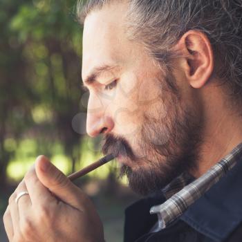 Bearded Asian man lights a cigar, outdoor square profile portrait with selective focus