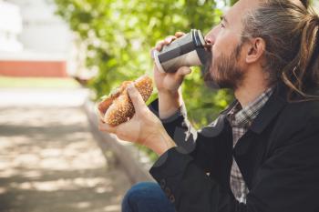 Bearded Asian man drinking coffee with hot dog  in summer park, outdoor profile portrait with selective focus