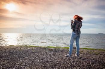 Teenage girl with red hair taking landscape phone photo on a seacoast in summer evening