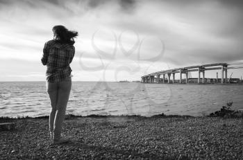 Girl stands on a sea coast, modern bridge under construction on a horizon, black and white photo