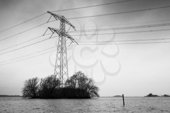 Transmission power tower, electricity pylon stands on small island with bare trees. Steel lattice tower, used to support an overhead power line. Black and white photo