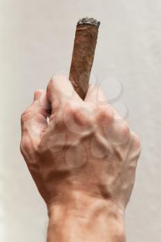 Gesture with handmade cigar in male hand, close-up photo with selective focus over white wall background 
