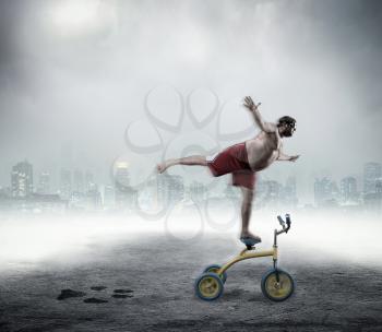 Nerdy adult man standing on a small bicycle on gray background