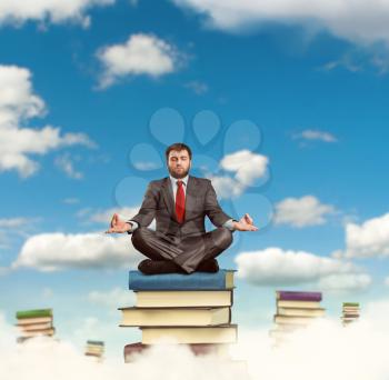 Businessman meditates sitting on the heap of the books in the clouds