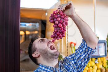 Portrait of hungry man in shirt trying to eat a bunch of grapes in supermarket. Boxes with fruits and vegetables on the background. 
