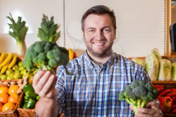 Smiling man holding broccoli at arm's length. Supermarket on the background.