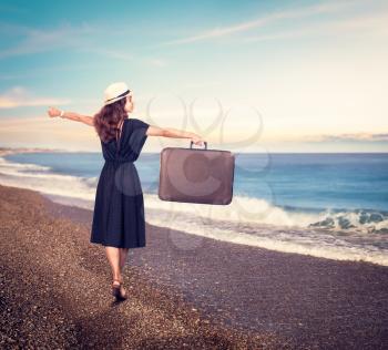 Young beautiful woman in black dress and white hat with suitcase on the beach. 