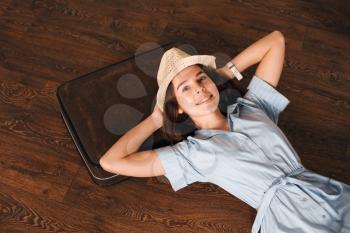 Smiling girl in light blue dress and white hat put her head on suitcase and dreams of a future journey. Girl lies on wooden floor. Travel waiting concept.