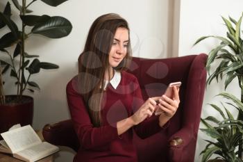 Young woman sitting in a red armchair with cell phone. Opened book and home plants on the background.