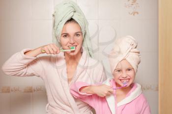 Happy mother and little daughter with toothbrushes teaching to brush their teeth in bathroom.