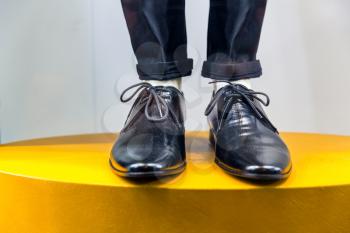 Mannequin legs in black classic shoes and folded trousers standing on yellow pedestal. White background.
