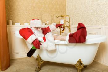 Funny drunk Santa Claus with alcohol bottle in hand sleeping in a bath. Christmas humor