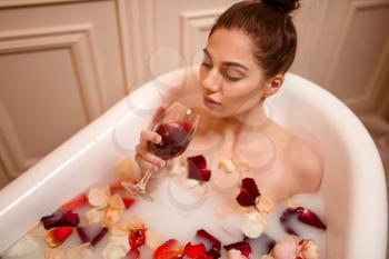 Beautiful nude woman holding a glass of red wine in bath with rose petals and foam.