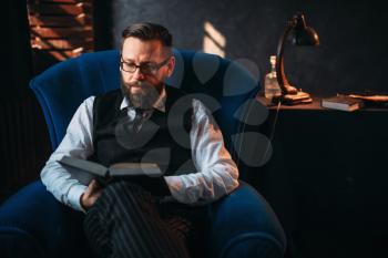 Seriuos bearded writer in glasses reads handwritten text against the window with sunlight. Retro typewriter, feather, crystal decanter, books and vintage lamp on the desk