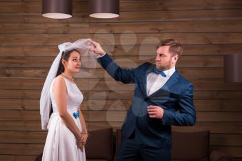 Young bride in white dress and veil against serious groom in suit, wooden background