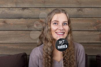 Young smiling woman holding funny icon on a stick with  just married inscription, wooden background. Fun photo props and accessories for shoots