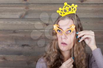 Young woman with funny glasses and crown on a sticks, wooden background. Fun photo props and accessories for shoots