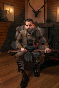 Hunter man with old gun in vintage traditional hunting clothing standing against antique chest. Fireplace, stuffed wild animals, bear skin and other trophies on background