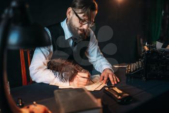 Bearded journalistr in glasses writes with a feather. Retro typewriter, books and vintage lamp on the desk