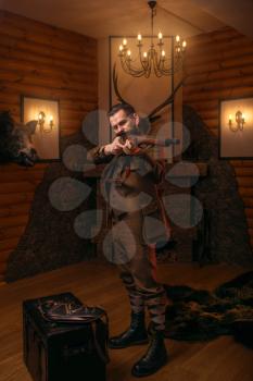 Senior hunter in retro traditional hunting clothing aims of the antique shotgun against old chest. Fireplace, stuffed wild animals, bear skin and other trophies on background