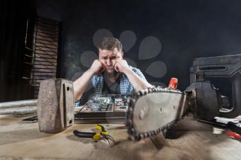 Engineer repairs laptop, repairman fix problem with electronic components. Chainsaw and anvil on the table, engineering humor