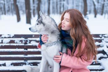 Happy woman sitting on the bench with siberian husky, snowy forest on background. Cute girl hugs with charming dog. Real friendship with pet