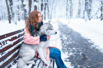Happy woman sitting on the bench with siberian husky, snowy forest on background. Cute girl hugs with charming dog. Real friendship with pet