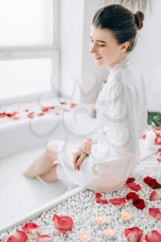 Elegant woman in white bathrobe sitting on the edge of the bath decorated with rose petals. Luxury bathroom interior
