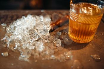 Brown beverage in a glass and ice on bar counter closeup, nobody. Refreshing of alcoholic drink, cold crystals on the table