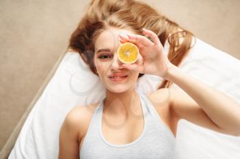 Young woman in underwear lies in bed and looks through a slice of lemon. Girl wake up in the morning in bedroom