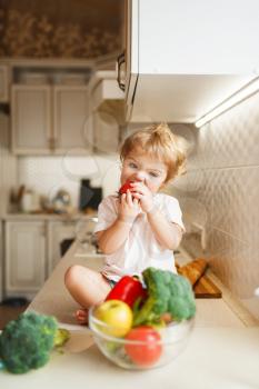 Little girl sitting on the table and eats tomato. Female baby tasting vegetables from the bowl on the kitchen. Child tastes vegetarian food