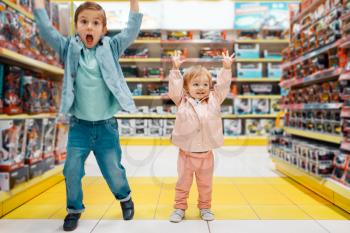 Little boy and girl raised their hands up at the shelf in kids store, happy children. Brother and sister choosing toys in supermarket, family shopping, young customers