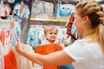 Mother with her little girl choosing clothes in kids store. Mom and child buying dress in supermarket together