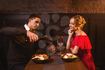 Man pours wine into a glass, young love couple in restaurant, romantic date. Elegant woman in red dress and her man, anniversary celebration