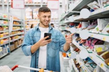 Man with cart makes a purchase by the list on phone in supermarket. Male customer in shop, husband with trolley choosing consumer goods, family shopping