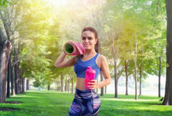 Athletic girl with sport bottle and yoga or fitness mat in summer park. Woman on morning fit workout