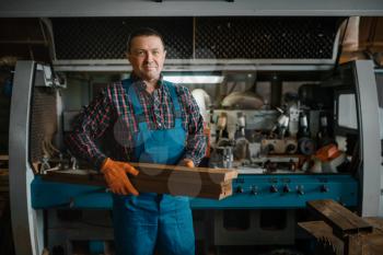 Carpenter in uniform holds boards, woodworking machine on background, lumber industry, carpentry. Wood processing on factory, forest sawing in lumberyard