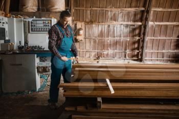 Carpenter with measuring tape measures boards, woodworking machine on background, lumber industry, carpentry. Wood processing on factory, forest sawing in lumberyard